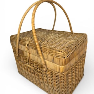 Op de afbeelding: Een rechthoekige picknickmand van riet met een scharnierend deksel en twee gebogen handvatten. De mand is geweven met een lichtbruin materiaal en heeft een sluiting. Het ontwerp is klassiek.