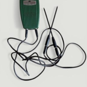 Op de afbeelding: Een vintage groen elektronisch apparaat met een geribbeld oppervlak en zwarte draden. Het apparaat heeft een zilveren label met tekst. Twee zwarte staven zijn gekruist, verbonden met het apparaat door zwarte draden.
