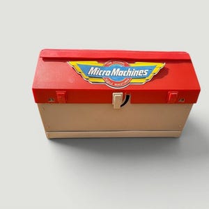 May include: A vintage Micro Machines toy box in red and beige. The box features the Micro Machines logo in yellow, blue, and white. The box has a rectangular shape with a hinged lid and a white latch.