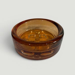 May include: A round, amber-colored glass ashtray. The ashtray has a thick, rounded rim and a decorative pattern around the sides. The interior features a cross-shaped design.