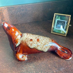 May include: A brown and white ceramic seal figurine with a speckled glaze. The seal is lying on its belly with its head turned to the side.