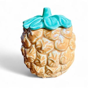May include: A ceramic pineapple-shaped jar with a green lid. The jar is decorated with a textured pineapple design.
