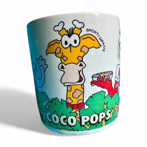 May include: White ceramic mug featuring a cartoon giraffe with the text "Shorty Giraffe" and "Coco Pops". The giraffe is yellow with brown spots and a red bow tie. Other cartoon characters are also visible.