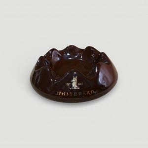 May include: A glossy, dark brown ashtray with a wavy rim. The ashtray has a circular shape and features the text "WHITEBREAD" in gold lettering, along with a small emblem and the year "1742".