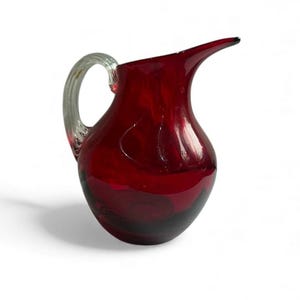May include: A ruby red glass pitcher with a clear, ribbed handle and a spout. The pitcher has a rounded body and a dark base. The glass appears to have a glossy finish, reflecting light. The pitcher is likely used for serving beverages.