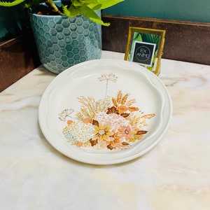 Peut inclure: Une assiette en céramique blanche avec un motif floral composé de fleurs brunes, orange et bleues et d'épis de blé. L'assiette a un bord festonné.