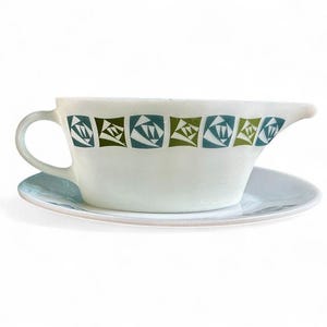 Puede incluir: Una salsera de cerámica blanca con su platillo a juego. La salsera tiene un asa y un pico vertedor. Está decorada con un patrón repetitivo de formas geométricas verdes y azules. El platillo es de color azul claro.