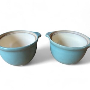 May include: A pair of light blue ceramic bowls, each with a handle on either side. The bowls have a rounded shape with a white interior and a light brown rim. Suitable for serving soups or other dishes.