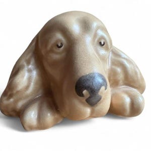 Peut inclure: Figurine de chien en céramique marron clair, avec un visage détaillé, des yeux sombres et un nez noir. Le chien a de longues oreilles tombantes et des pattes visibles. Un objet décoratif pour les amoureux des chiens.