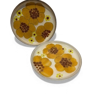 Op de afbeelding: Twee ronde keramische borden met een bloemenontwerp. De borden hebben grote gele bloemen met bruine centra en kleinere gele bloemen op een crèmekleurige achtergrond. De randen zijn bruin.