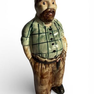 Puede incluir: Figura de cerámica de un hombre con barba, con una camisa a cuadros verde y pantalones marrones. La figura tiene zapatos negros y las manos en los bolsillos. La figura está de pie.