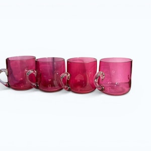 May include: Four ruby-red glass mugs with handles are displayed in a line. The mugs have a rounded form and a smooth surface. The handles are also glass and attached to the side of each mug. The mugs are set against a white backdrop.