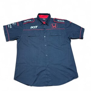 Puede incluir: Camisa azul marino de manga corta con ribetes y detalles en rojo. La camisa presenta el logotipo de Honda y la palabra "acer" en blanco. Tiene dos bolsillos en el pecho y cierre de botones.