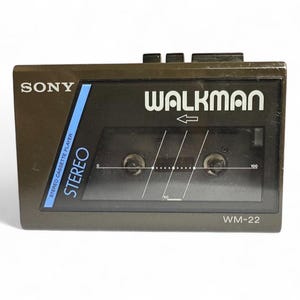 May include: A vintage Sony Walkman WM-22 stereo cassette player. The device is dark gray with the word "WALKMAN" in white. The cassette window shows the tape and a white graphic. The word "SONY" is at the top left.