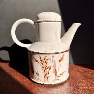 May include: A white ceramic teapot with a brown wheat design. The teapot has a round body and a spout on one side. The lid is also white and has a brown rim.