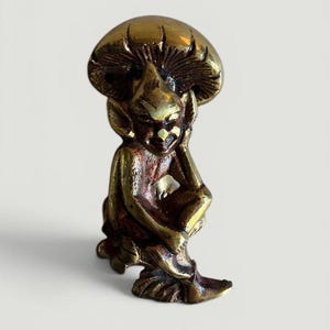 May include: A brass figurine of a seated gnome with a mushroom cap. The gnome is detailed with pointed ears and a smiling expression. The mushroom cap provides a canopy over the gnome's head. The figurine has a vintage, antique appearance.