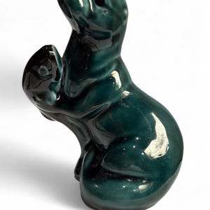 Puede incluir: Figurita de cerámica verde azulado de una foca con una cría. La escultura brillante representa a la foca adulta mirando hacia arriba, sosteniendo una foca más pequeña. La figura tiene una superficie lisa y reflectante.