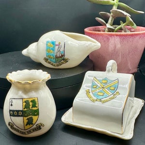 May include: Three small ceramic containers with gold trim and painted crests. The containers are shaped like a seashell, a pouch, and a house. The crests feature different designs and text, including "FRINSTON ON SEA", "WELLINGTON", and "SOMERSET".