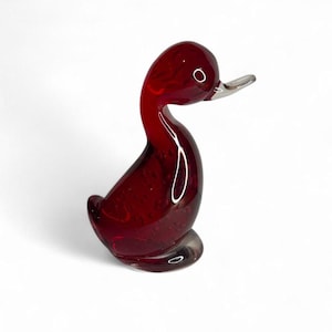 Puede incluir: Figura de pato de cristal rojo rubí con pico plateado. El pato está en posición sentada con la cabeza ligeramente girada hacia un lado. El cristal tiene un acabado brillante y es transparente.