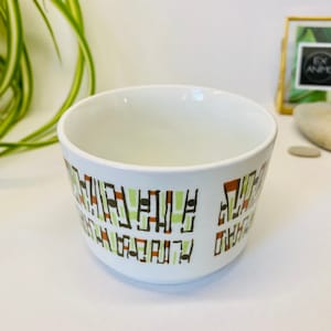 May include: White ceramic bowl with a geometric pattern in brown, green, and orange. The pattern is abstract and resembles a stylized cityscape.