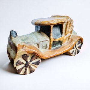 May include: A ceramic sculpture of a vintage car with brown, blue, and white glaze. The car has a detailed design with a rounded roof and a large front wheel.