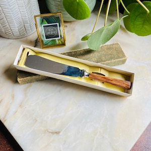 May include: A silver butter knife with a wooden handle in a white box with a green and gold patterned box underneath.