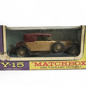 Może przedstawiać: Zabytkowy model samochodu Matchbox Y-15 1930 Packard Victoria w oryginalnym pudełku. Samochód jest złoty z bordowym dachem kabrioletu i czarnymi oponami. Pudełko jest fioletowo-żółte z logo Matchbox.