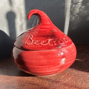 Könnte beinhalten: Ein roter Keramikbehälter in Form einer Roten Bete mit Deckel. Der Deckel hat einen Griff und das Wort "Beetroot" ist darauf geschrieben.
