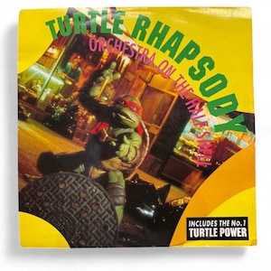 Puede incluir: Una carátula de CD amarilla con el texto "Turtle Rhapsody" y "Orchestra on the Half Shell". La portada presenta un personaje de las Tortugas Ninja con una máscara roja, con una alcantarilla en primer plano. También es visible el texto "Includes the No. 1 Turtle Power".