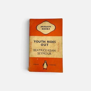 Puede incluir: Un libro de bolsillo vintage de Penguin Books titulado Youth Rides Out de Beatrice Kean Seymour. La portada presenta un esquema de colores naranja brillante y crema con el clásico logotipo del pingüino en la parte inferior, texto negro y la palabra FICTION impresa verticalmente.