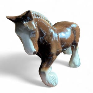 May include: A ceramic horse figurine in shades of brown, white, and tan. The horse has a glossy finish and is posed with its head down. The figurine is a decorative item, likely for display.