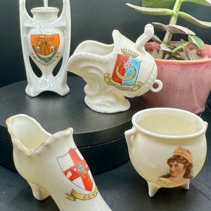 May include: Four small white ceramic figurines with painted details. Each figurine depicts a different coat of arms with the city name below. The figurines are on a black surface.