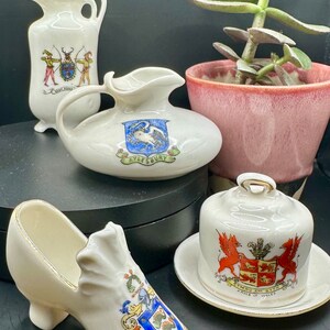 Puede incluir: Cuatro jarras en miniatura de cerámica blanca con ribetes dorados y escudos de armas pintados. Las jarras tienen forma de jarra, zapato, cúpula y jarra tradicional. Las jarras están decoradas con escudos de armas coloridos y texto.