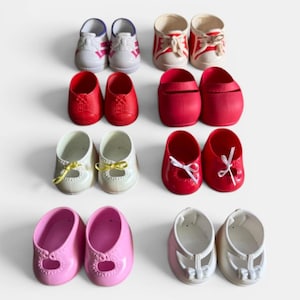 Op de afbeelding: Een verzameling poppenschoenen in verschillende stijlen en kleuren, waaronder rood, roze, wit en crème. De schoenen hebben details zoals veters, strikken en uitgesneden ontwerpen. Sommige zijn sneakers, andere Mary Janes.