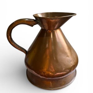 May include: A copper jug with a curved handle and a flared spout. The jug has a bulbous body and a wide, circular base. The copper has a warm, reddish-brown hue and a polished finish. The jug is likely used for pouring liquids.