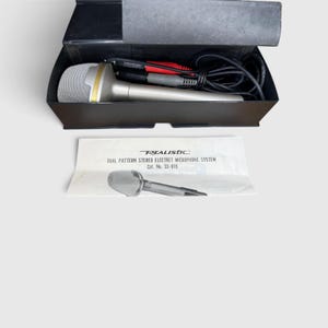 Op de afbeelding: Een zilverkleurig Realistic dual pattern stereo electret microfoonsysteem met een zwarte kabel en rood-zwarte connectoren, opgeborgen in een zwarte koffer. Een witte handleiding met de tekst Realistic Dual Pattern Stereo Electret Microphone System Cat. No. 33-919 is inbegrepen.