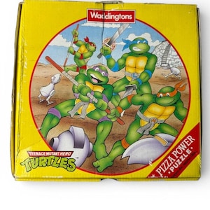 May include: A yellow box featuring the Teenage Mutant Ninja Turtles. The box art shows the four turtles in action, with weapons, inside a red circle. The words "Pizza Power Puzzle" are on the box.