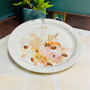 Peut inclure: Une assiette plate en céramique blanche avec un motif floral représentant un bouquet de fleurs brunes, jaunes et roses avec des feuilles vertes et des épis de blé. L'assiette a un bord légèrement festonné.