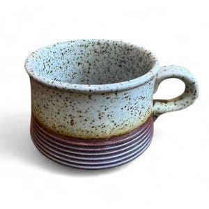 May include: A ceramic mug with a speckled, light blue and white exterior. The base has a brown and purple striped design. The mug has a rounded handle. Suitable for hot or cold drinks. The mug is approximately 10 cm tall.