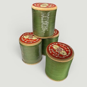 May include: Four spools of green thread are stacked. Each spool has a wooden base and a red and gold label. The labels read "No. 60 3 Cord" and "3 Ounce". The thread is wrapped in clear plastic.