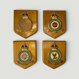 Könnte beinhalten: Vier hölzerne Wandplaketten mit militärischen Wappen. Jede schildförmige Plakette ist mit einer goldfarbenen Krone versehen. Die Wappen enthalten Texte wie JE VOLE A TOUS VENTS, WINGS IN THE SUN, EQUITES NOSTRI ARMA SUMUNT und SUITRA MARI SUITRA TERRAMQUE.