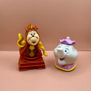 May include: A brown and yellow clock figurine with a smiling face and a purple and white teapot figurine with a smiling face. Both figurines are from the Disney movie Beauty and the Beast.