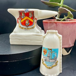 May include: Two porcelain figurines, one shaped like an anvil with a coat of arms featuring three figures and the text "Fiat Voluntas Dei Odiham", the other shaped like a bell with a coat of arms featuring a ship and the text "Lydd".