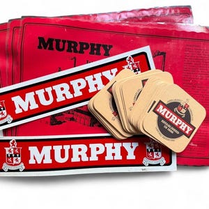 Op de afbeelding: Een verzameling rode en witte Murphy's bierartikelen. De afbeelding toont rode placemats, rechthoekige stickers en vierkante onderzetters. Elk item heeft het Murphy's logo, een wapen en de tekst "ESTABLISHED IN 1856".