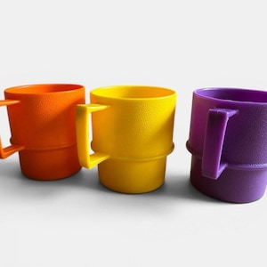 Puede incluir: Tres tazas de plástico de colores naranja, amarillo y morado. Cada taza tiene una superficie texturizada, un cuerpo cilíndrico y un asa cuadrada. Las tazas están dispuestas una al lado de la otra sobre un fondo blanco.