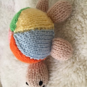 May include: A colorful knitted turtle toy with a patchwork shell in shades of pink, orange, yellow, green, and blue. The turtle has a pink body and a small black nose.