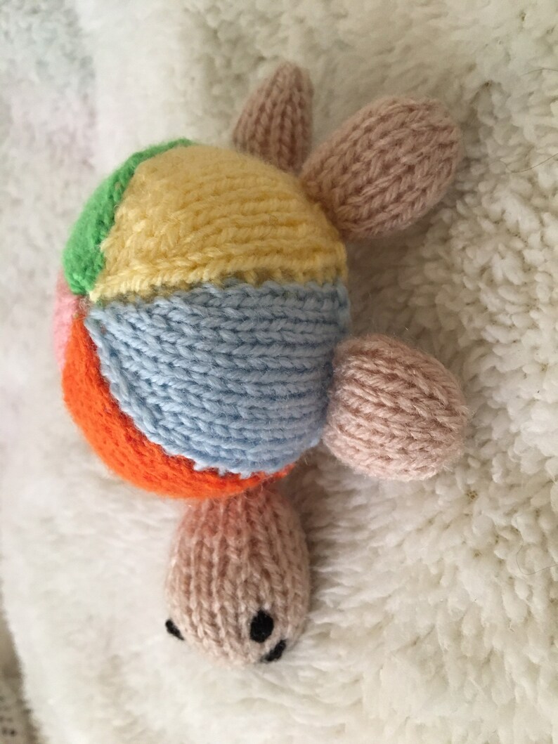 May include: A colorful knitted turtle toy with a patchwork shell in shades of pink, orange, yellow, green, and blue. The turtle has a pink body and a small black nose.