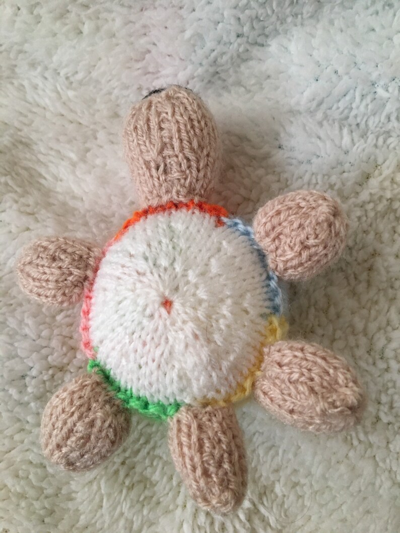 May include: A knitted turtle toy with a white shell and colorful accents. The turtle has a brown body and a black nose.