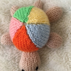 May include: A knitted turtle toy with a multi-colored shell. The shell is made of six different colored panels: green, yellow, pink, orange, blue, and white. The turtle has a light brown body and a small, brown nose.