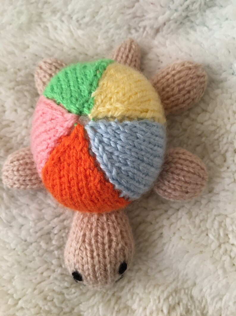 May include: A knitted turtle toy with a multi-colored shell. The shell is made of six different colored panels: green, yellow, pink, orange, blue, and white. The turtle has a light brown body and a small, brown nose.